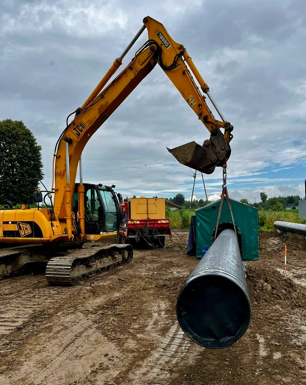 Koparka JCB przenosząca ciężką rurę stalową na placu budowy infrastruktury przesyłowej.
