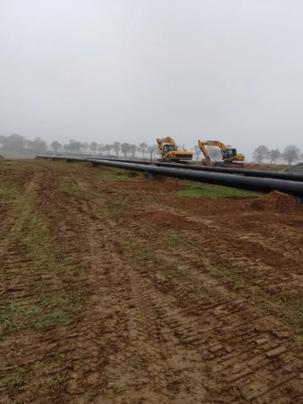 Długie odcinki zespawanych rur przygotowane do ułożenia w terenie, w tle pracujący sprzęt ciężki.